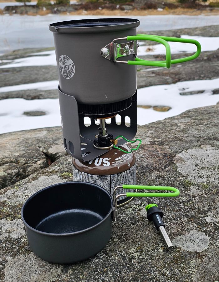 Ett grått Optimus Crux stormkök med gröna handtag står monterat på en primus vintergas. En liten stekpanna står framför och en piezotändare ligger bredvid. I bakgrunden syns klippor och en frusen sjön.