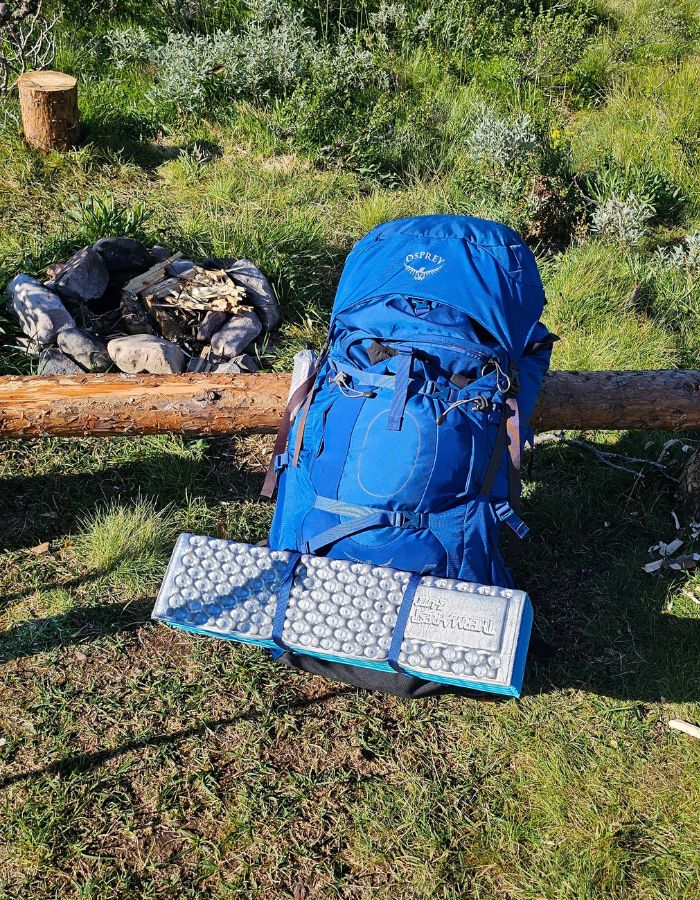 Ett Therm-A-Rest Z-Lite Sol cellplast liggunderlag fäst på en vandringsryggsäck från Osprey. Ryggsäcken står lutad mot en stock med en eldstad i bakgrunden.