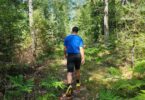 Man går i skogen iklädd vandringsshorts i grått och blå t-shirt. I bakgrunden syns gröna barrträd.