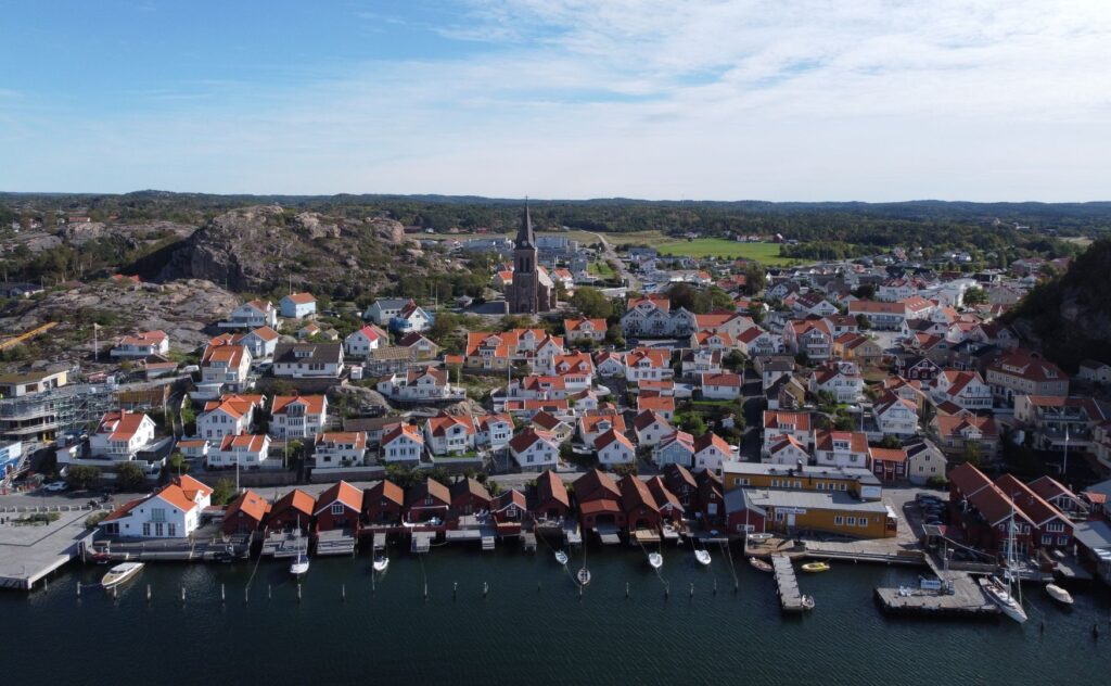 En drönarbild på FJällbacka tagen från havet. Röda sjöbodar kantar bryggan och vita hus klättrar upp på klipporna.