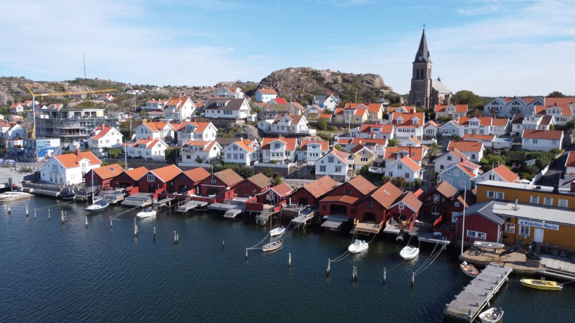 En drönarbild på FJällbacka tagen från havet. Röda sjöbodar kantar bryggan och vita hus klättrar upp på klipporna.
