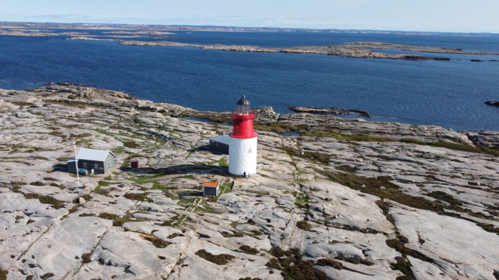 Drönarbild på Hållö med den röd-vita Hållöfyren mitt i bild. Karga klippor med det blå havet i bakgrunden.