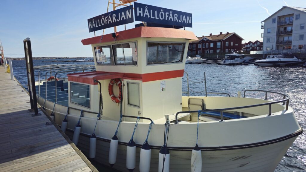 Hållöfärjan anlagd vid Smögenbryggan. En stor, öppen, vit plastbåt med skylt på taket ligger vid en träbrygga.