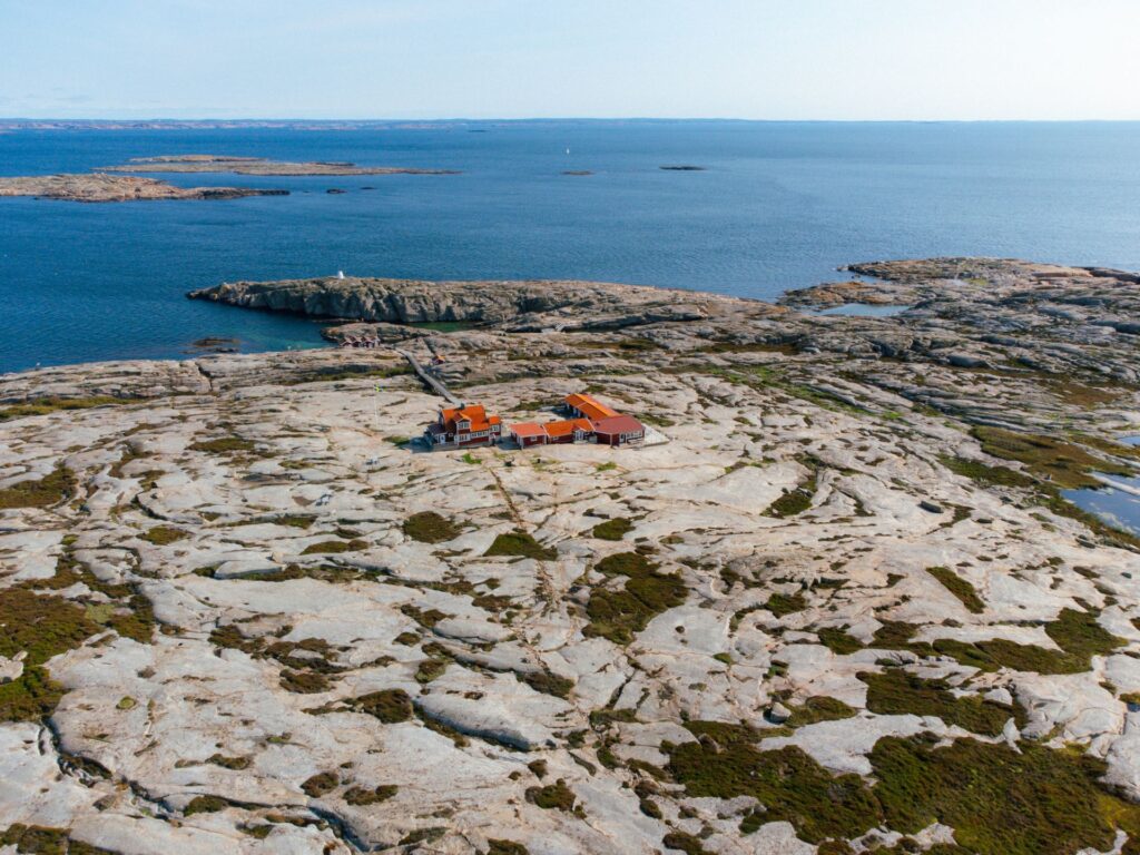Drönarbild av Hållös steniga landskap. Man kan se Utpost Hållös röda stugor och mot horizonten det blå havet.